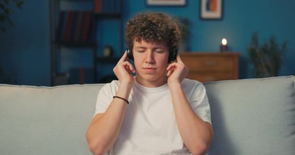 Un adolescente con el pelo rizado se sienta en un sofá de la sala de estar con auriculares inalámbricos — Vídeos de Stock