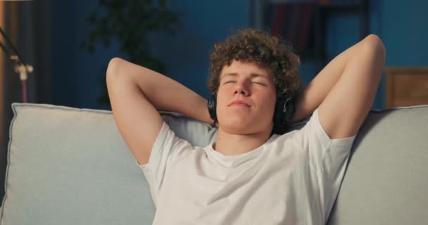 A férfi a kanapén pihen, kezeket tarkóra téve, zenét hallgat, podcastet játszik. — Stock videók