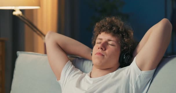 Ένας νεαρός αναπαύεται σε έναν καναπέ, τα χέρια πίσω από το κεφάλι, ακούγοντας μουσική. — Αρχείο Βίντεο