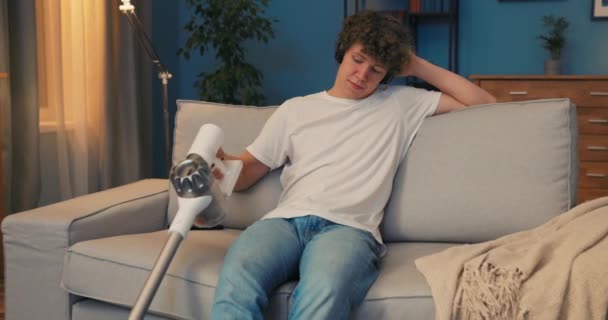 Мальчик принудительно пылесосит в комнате по вечерам, сидя на диване — стоковое видео