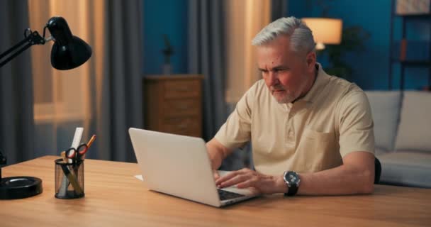 Anciano sentado en el escritorio con portátil por la noche. Senior trabaja en un salón. — Vídeos de Stock