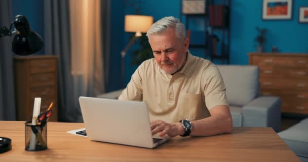 Elderly man sitting at desk with laptop in the evening. Grandfather talks to grandchildren online. — Stock Video