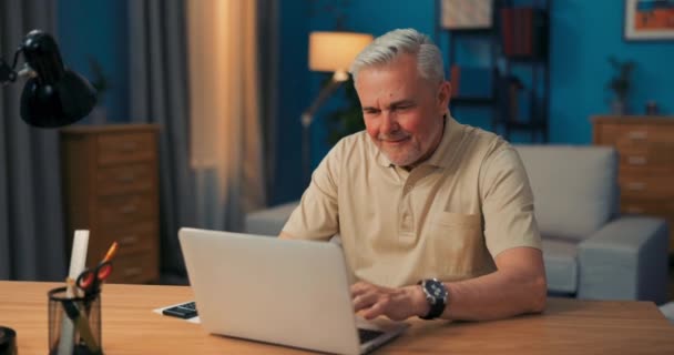Idős férfi ül az asztalnál laptoppal este. Nagyapa online beszél az unokáival. A... — Stock videók