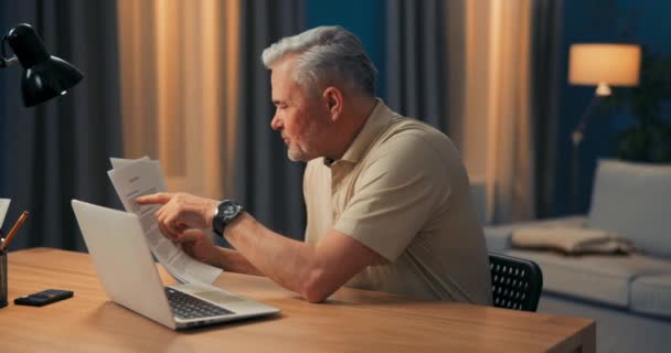 Mannen sitter framför datorn i vardagsrummet på kvällen. En äldre man sitter — Stockvideo