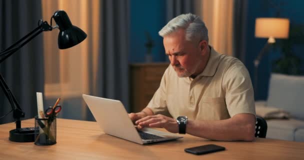 Пожилой человек с седыми волосами работает на ноутбуке за столом и лампой вечером. Зрелый — стоковое видео
