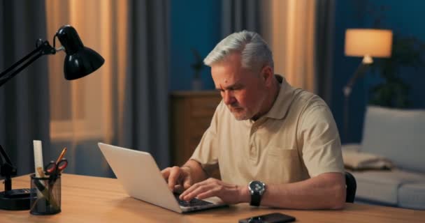 Пожилой человек с седыми волосами работает на ноутбуке за столом и лампой вечером. Зрелый — стоковое видео