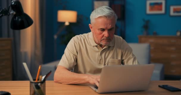 Äldre man sitter vid skrivbordet med laptop på kvällen. Senior arbetar i en salong. — Stockvideo