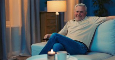 Gri saçlı yaşlı bir adam oturma odasında rahat bir şekilde oturuyor.