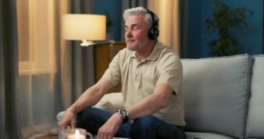 Yaşlı bir adam koltukta rahat bir pozisyonda oturuyor, kablosuz kulaklık takıyor.,