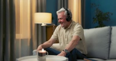 Yaşlı bir adam koltukta rahat bir pozisyonda oturur, kablosuz kulaklıkları takar, dinler.