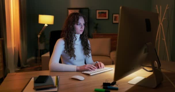 Красивая молодая девушка с кудрявыми волосами сидит вечером перед экраном компьютера, — стоковое видео