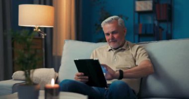 Oturma odasında tabletle dinlenen gülümseyen gri saçlı adam. Emekli adam haberleri okuyor.