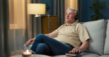 Oturma odasındaki kanepede oturan yaşlı bir adam müzik dinliyor.