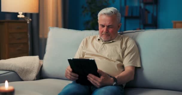 Улыбающийся седой мужчина отдыхает в гостиной на диване с планшетом. Пенсионер читает новости — стоковое видео