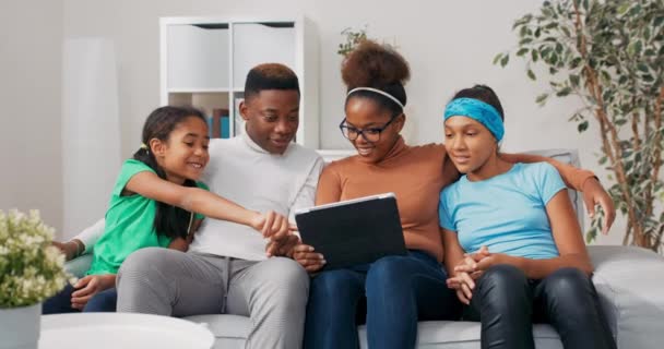 Família feliz sentado no sofá em um apartamento recém-comprado um casal e suas duas filhas navegar sites de móveis em um tablet, planejar uma renovação, a menina escolhe mobiliário — Vídeo de Stock