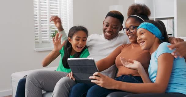 Uma família feliz senta-se no sofá, relaxando, passando o tempo juntos à tarde, pais com duas filhas acenando para a câmera do tablet, tendo uma conversa em vídeo com amigos — Vídeo de Stock