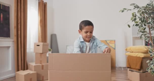 Um menino bonito desempacota sua caixa de brinquedos depois de se mudar para uma nova casa, puxa — Vídeo de Stock