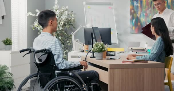 En man sitter i rullstol vid ett skrivbord på ett kontor mitt emot honom. — Stockvideo