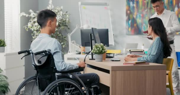 Un hombre se sienta en una silla de ruedas en un escritorio en una oficina, frente a él — Vídeos de Stock