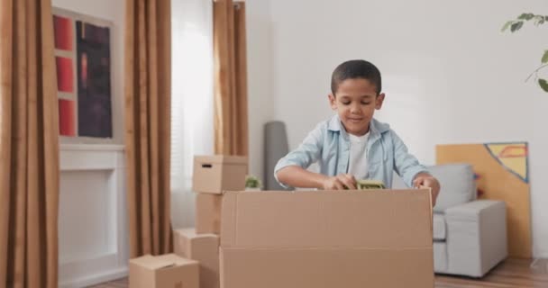 Um menino bonito desempacota sua caixa de brinquedos depois de se mudar para uma nova casa, puxa — Vídeo de Stock