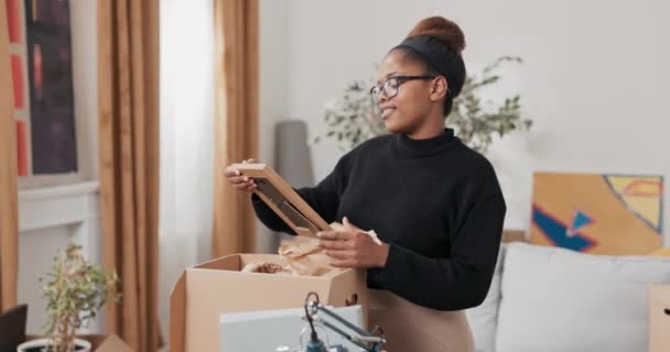 Finding souvenirs during a move, a woman in glasses unpacks a cardboard box in which — Stock Video