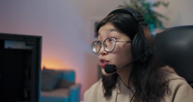 İnek gözlüklü bir kız bilgisayarda oyunlar oynuyor, oyuncu kulaklıkları kulaklıklı rahat bir sandalyeye oturuyor, şok oluyor, sanal dünyayı yaşıyor, odada ışıklandırmaya öncülük ediyor.