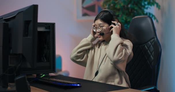 Kobieta dołącza do rozmowy z przyjaciółmi przez słuchawki wkłada grać w gry wideo dla wielu graczy. Dziewczyna jest zaawansowanym graczem mistrzostw e-sportu online. Spędza czas w pokoju oświetlony przez led — Wideo stockowe