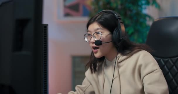 Профессиональная женщина-геймер готовится к онлайн-играм, надевает наушники, разговаривает со своими оппонентами, начинает раунд в игре, смотрит через очки на компьютерный монитор — стоковое видео