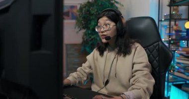 İnek gözlüklü kız bilgisayarda oyunlar oynuyor, profesyonel oyuncu kulaklıkları kulaklarında rahat bir koltukta oturuyor, şok oluyor, sanal dünyayı yaşıyor, odada ışıklandırmaya öncülük ediyor.