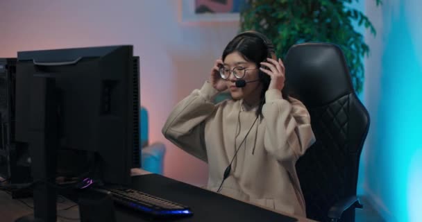 Beliebte Mädchen Streamer betritt Raum beleuchtet von LED-Lampen, setzt sich auf Stuhl vor Computer und setzt Headset auf, weibliche Gamer begrüßt ihre Fans, für die sie ein Online-Spiel live streamt — Stockvideo
