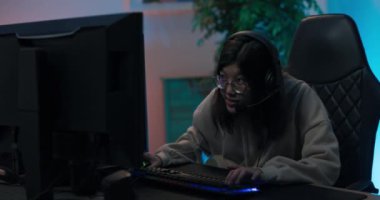 Gözlüklü kız bilgisayarda oynuyor, profesyonel oyuncu kulaklıkla rahat koltukta oturuyor, duygusal olarak duygulanmış, sanal dünyayı yaşıyor, odada ışık var.