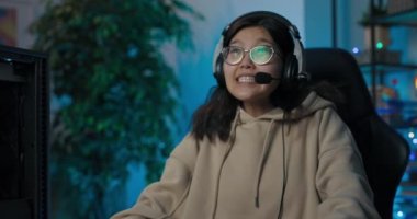 İnek gözlüklü genç Asyalı kız profesyonel oyuncu, kulaklık takıyor, karanlık bir odada bilgisayar oyunu oynuyor öncü neon ışıkları ile vücut hareketlerini engelleyen oyundaki duygulardan kaçınıyor.