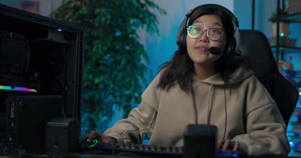 Chica sonriente sentada en una habitación frente a un monitor de computadora auriculares de equipos de juego profesionales con micrófono hablando con rivales luchando por la estrategia de victoria cuarto oscuro con luces led — Vídeo de stock