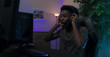 Profesyonel olarak bilgisayar oyunu oynayan gülümseyen adam koltuğunda rahatça oturup monitöre bakıyor, kulaklıkla takım üyeleriyle konuşuyor, oda neon mavisi.