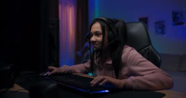 Kulaklık takmış, internetten video oyunu oynayan neşeli, profesyonel oyuncu kız. Arkadaşlarıyla sohbet ediyor. Renkli neon ışıkları odada.