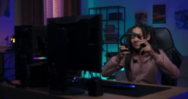 Gülümseyen kız gecelerini masa sandalyesinde geçiriyor kulaklığı takıyor, takım üyelerini selamlıyor, online arkadaşları, genç oyuncu kız sanal dünyada yaşıyor, oda ışıklarla aydınlatılıyor.