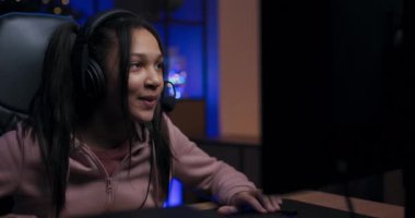 Odaklanmış oyuncunun monitörünün arkasından çekilmiş. Kızın oyun kulaklığı var, online oyunları kazanmak için strateji geliştiriyor. Gece karanlığında, ışıklarla aydınlatılmış bir oda.