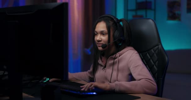 Joueuse pro en sweat-shirt rose est assis sur une chaise devant un ordinateur, la fille joue à des jeux informatiques avec une équipe à laquelle elle parle à travers un casque, elle regarde dans la caméra et sourit — Video