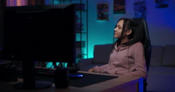 Jovem gamer menina senta-se na cadeira na frente do computador tem uma pausa de jogos descansa pensativo, pondera, sorri, ri, quarto à noite iluminado com luzes led coloridas — Vídeo de Stock