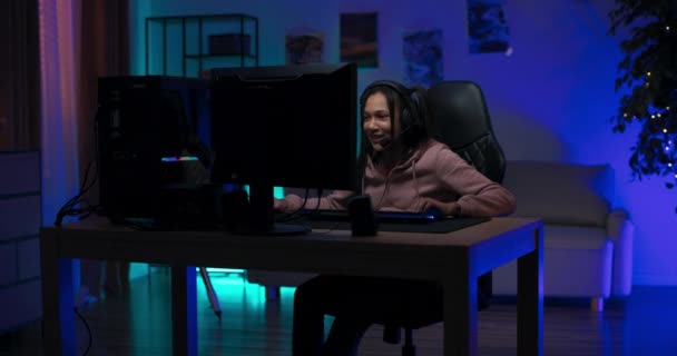 Радостная девушка геймер сидит перед компьютером в наушниках, профессиональное игровое оборудование, женщина выиграла раунд, прошла уровень, победный танец руками — стоковое видео