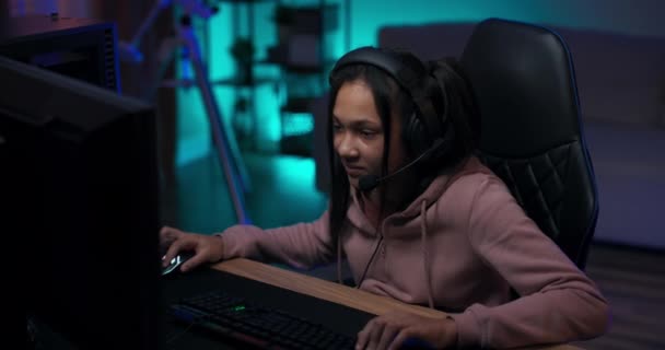 Дівчина-підліток захоплюється віртуальним світом грає стрільців на комп'ютері, молода дівчина енергійно клацає пальцями на клавіатурі, має навушники, аксесуари для підсвічування синьої кімнати — стокове відео