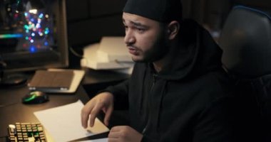 Kapüşonlu sweatshirt giyen yorgun, aşırı çalışan erkek programcı geceleri karanlık odada çalışıyor, kafası masanın üzerinde bilgisayarın önünde uyuyor, gözlerini ovuşturarak uyanıyor ve yazılım kodlamaya başlıyor.