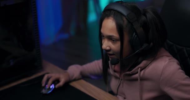Усміхнена молода дівчина сидить у кімнаті перед комп'ютером монітор професійне ігрове обладнання навушники з мікрофоном, розмовляючи з суперниками, борючись за стратегію перемоги темна кімната з світлодіодними ліхтарями — стокове відео