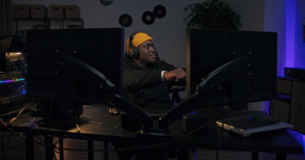 暗い部屋に導かれたライト男がコンピュータの前にスウェットシャツと黄色のキャップを着て座って点灯。音楽プロデューサースタジオラッパーリズムに手で仕事のダンスに満足曲を処理 — ストック動画