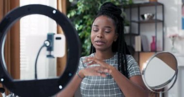 Etkilenen hayranlarını apartmandan karşılıyor, sosyal medya için makyaj ürünleri hakkında bir video yapıyor. Kadının önünde telefon için tripodu olan bir ayna ve halka lambası var. Uzaktan çalışma çevrimiçi