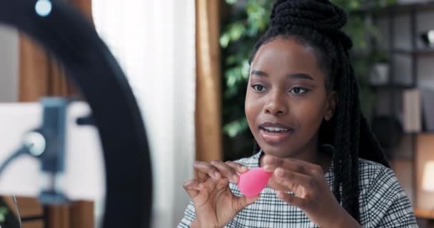 Maquiagem artista fornece treinamento remoto de casa. Mulher talentosa explica o princípio da esponja para aplicar a fundação, contornando produtos. Menina fala em telefone colocado no tripé com anel de luz — Vídeo de Stock