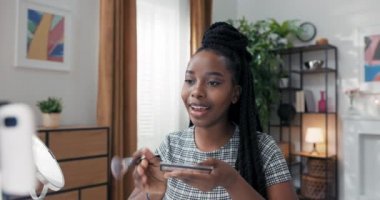 Bir kız telefon tripoduyla halka ışığı kullanarak video çekiyor. Makyaj sanatçısı yüz hatlarını nasıl düzelteceğine dair bir ders hazırlar. Yanağa brozer sürmek için fırça kullanır.