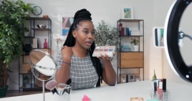 Gülümseyen esmer kadın güzellik blogcusu göz farı paleti gösteren ürünler öneriyor. Akıllı telefon ve halka ışığıyla video kaydediyor. Kadın makyaj sanatçısı etkileyicisi özel ders oluşturur