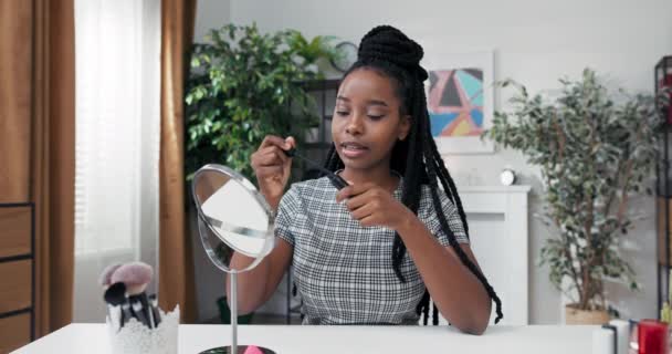 Maquiagem artista com dreadlocks e pele escura senta-se em pestanas de pintura de mesa enquanto olha em um espelho. O influenciador está gravando vlog, contando aos espectadores sobre os benefícios do rímel. — Vídeo de Stock