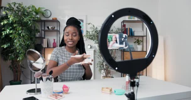 Artistul de machiaj experimentat de origine afro-americană aplică umbra ochilor pe pleoape, înregistrând un tutorial pe ochii afumați, în fața ei pe oglinda de masă, cosmetice și accesorii de frumusețe — Videoclip de stoc